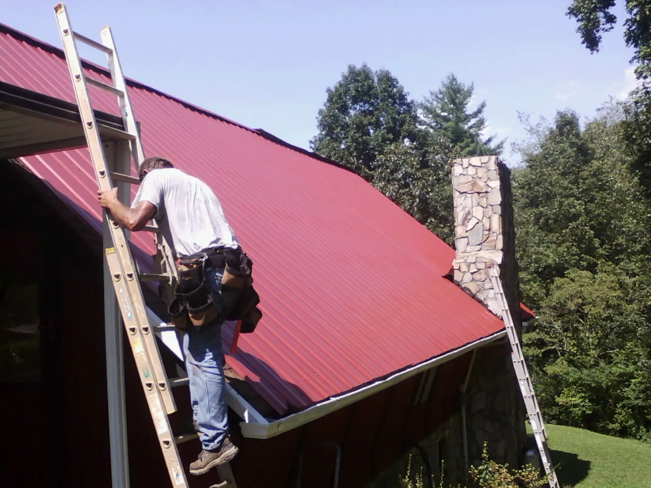 Roofer on a ladder beside a red metal roof and stone chimney — Soffit & Fascia Repair by Roofing Brothers in Carbonado